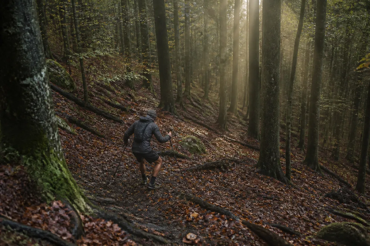Coureur de trail en descente sur un sentier forestier, bâtons plantés en appui devant lui